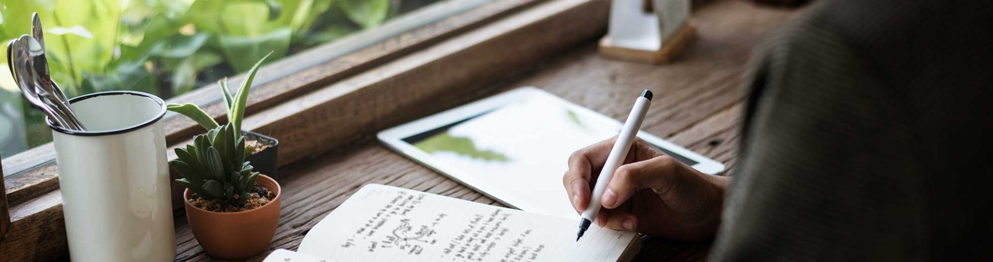 Une main tient un stylo juste au dessus de feuillets annotés posées sur un bureau devant une fenêtre.
